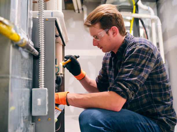 HVAC technician wearing safety glasses and gloves crouching to inspect a furnace unit with a flashlight in a utility room.