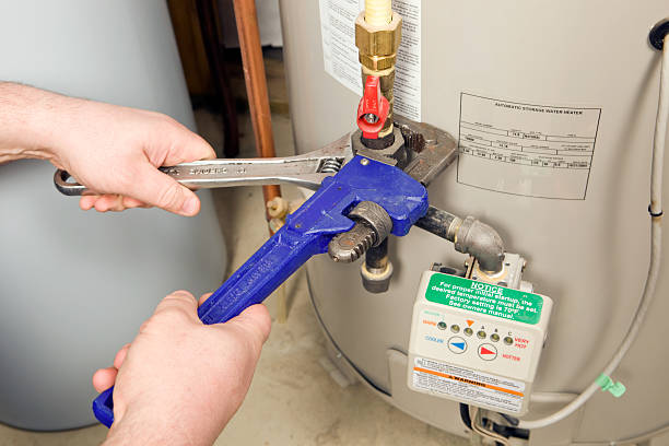 Two hands using pipe wrenches to tighten fittings on a gas storage water heater with a temperature control