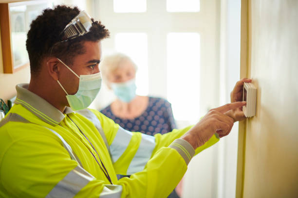Technician in hi-vis jacket and face mask adjusting a wall thermostat while an elderly customer looks on